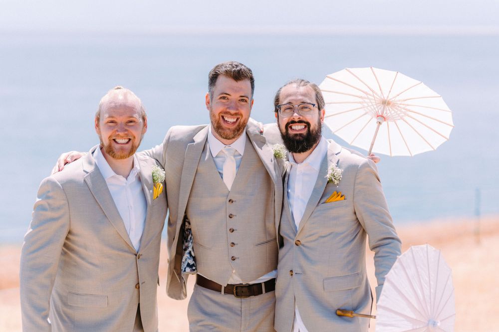 Three people in light colored suits smile while posing with parasols against blue sky.