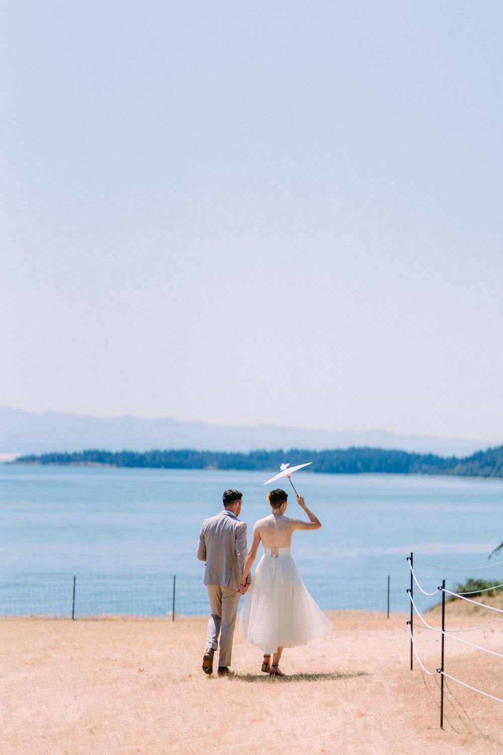 Couple walks along coastal path holding white parasol against ocean backdrop.