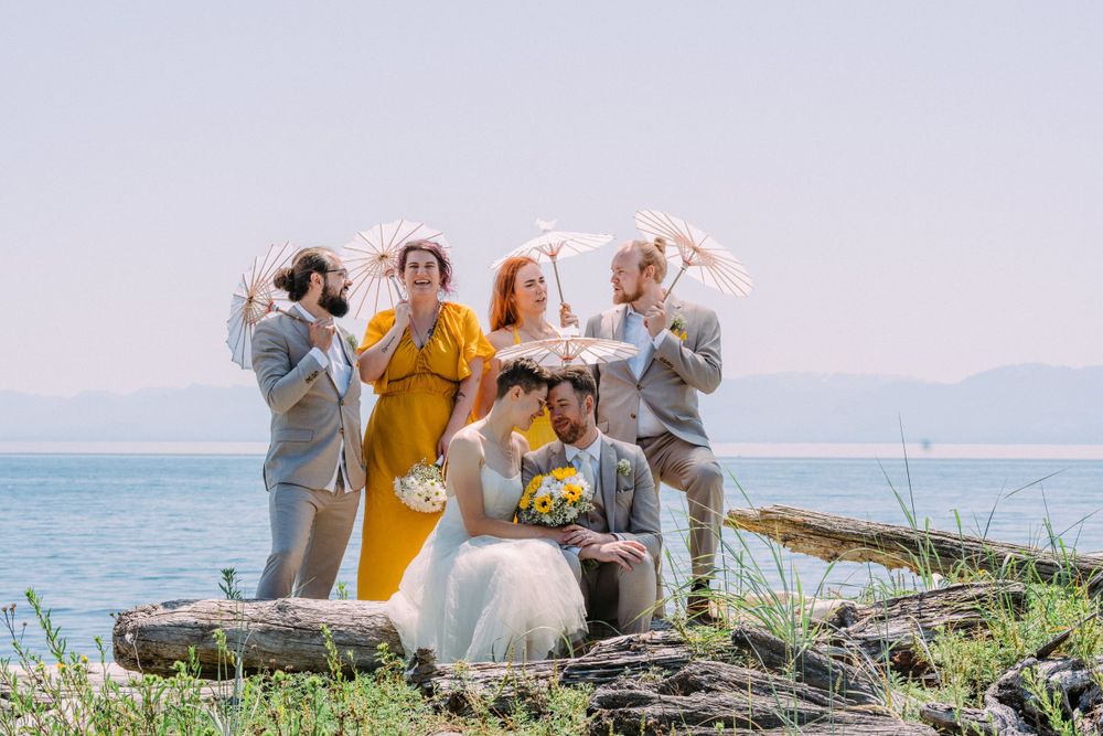 Wedding group poses playfully with parasols on driftwood by ocean.