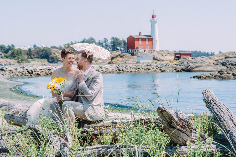 A wedding couple embraces on rocky shoreline with red lighthouse in background at seaside location.