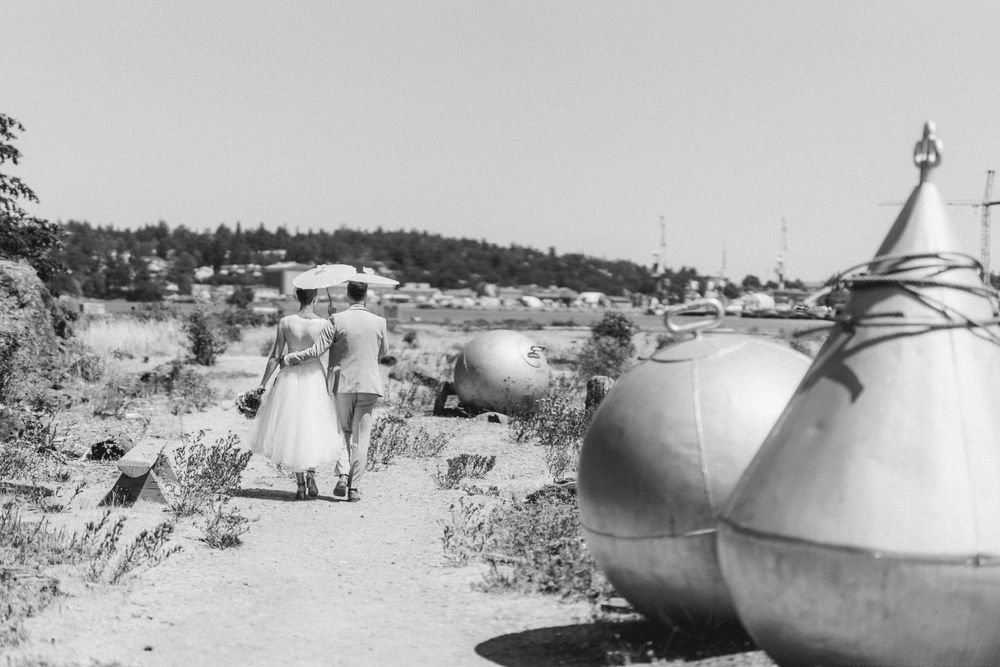 Vintage black and white photo of two people walking on rocky path near lighthouse and vessels.