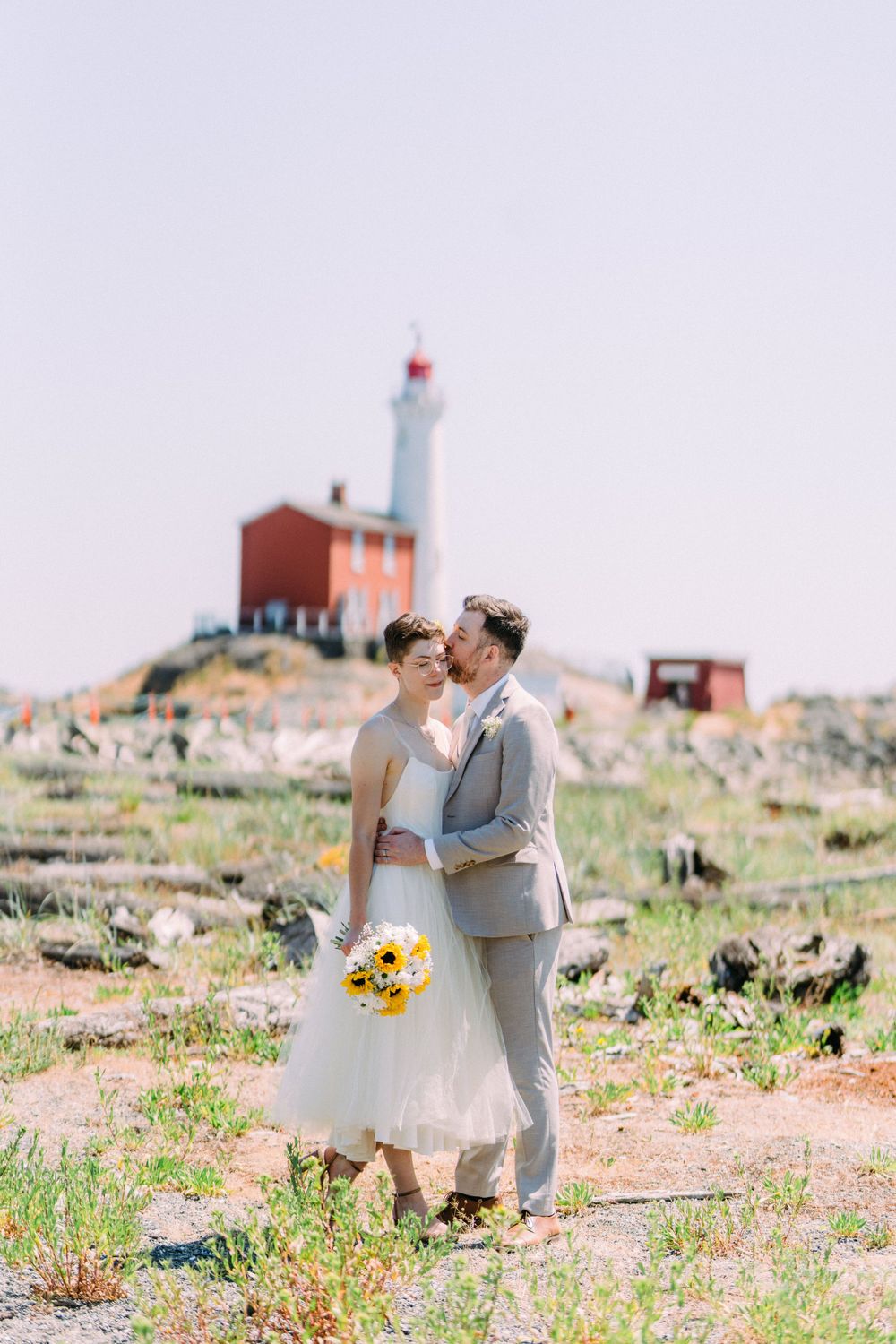A romantic wedding photo with couple on rocky terrain with lighthouse and bright yellow bouquet.
