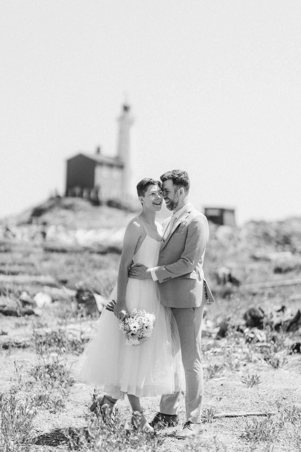 Black and white wedding portrait with lighthouse backdrop on rocky coastal landscape.