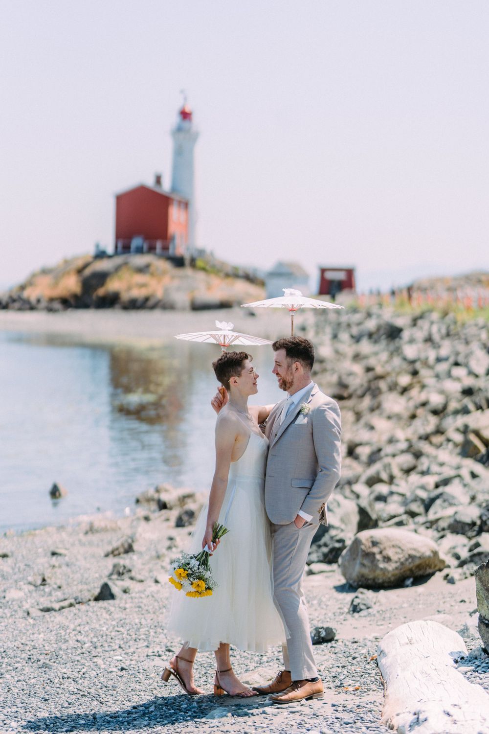Coastal wedding scene with couple standing on rocky beach near red and white lighthouse.