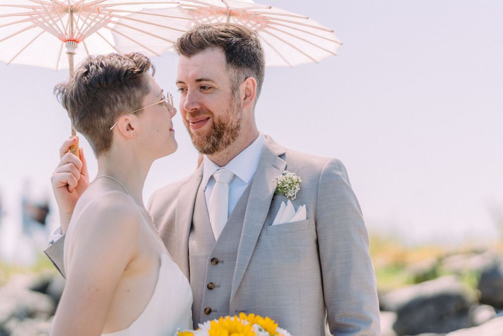 Wedding portrait featuring white parasol and yellow sunflower bouquet against soft sky.