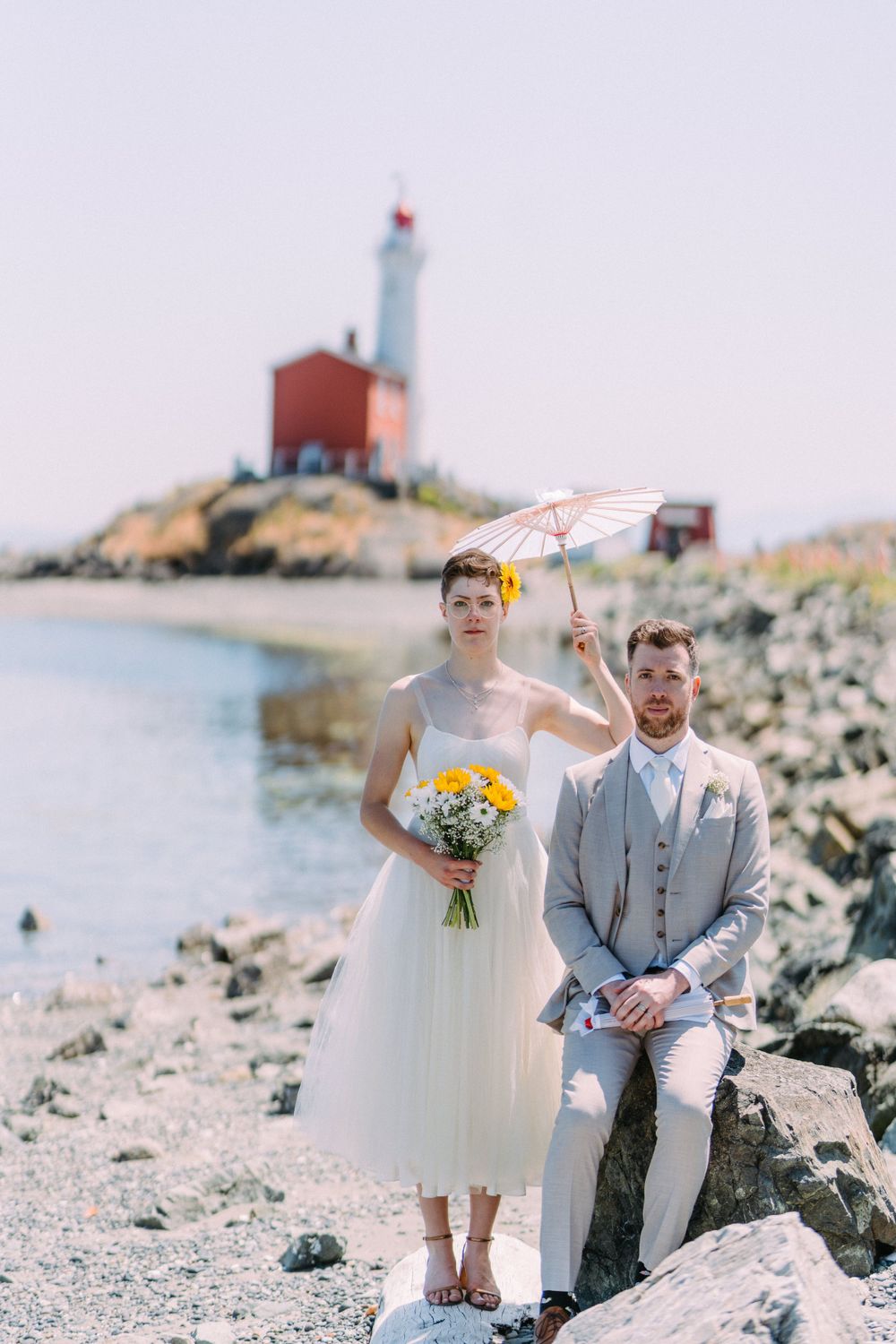Romantic wedding photo with parasol and lighthouse featuring tea-length dress and grey suit.