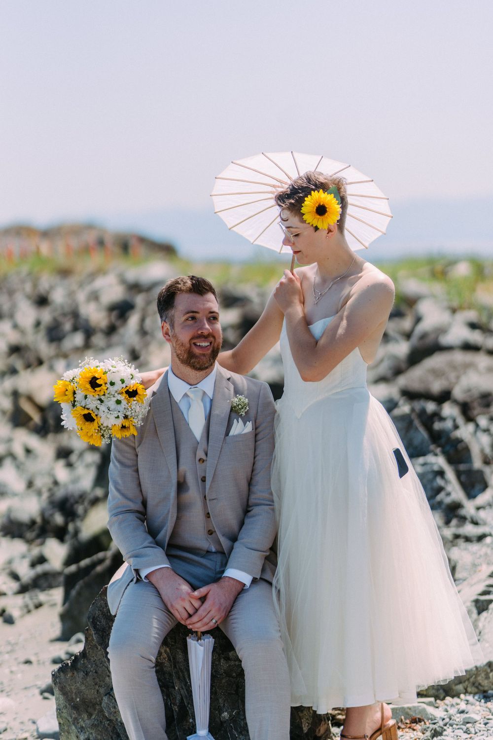 Playful wedding portrait on rocky coast with parasol and sunflower bouquet.