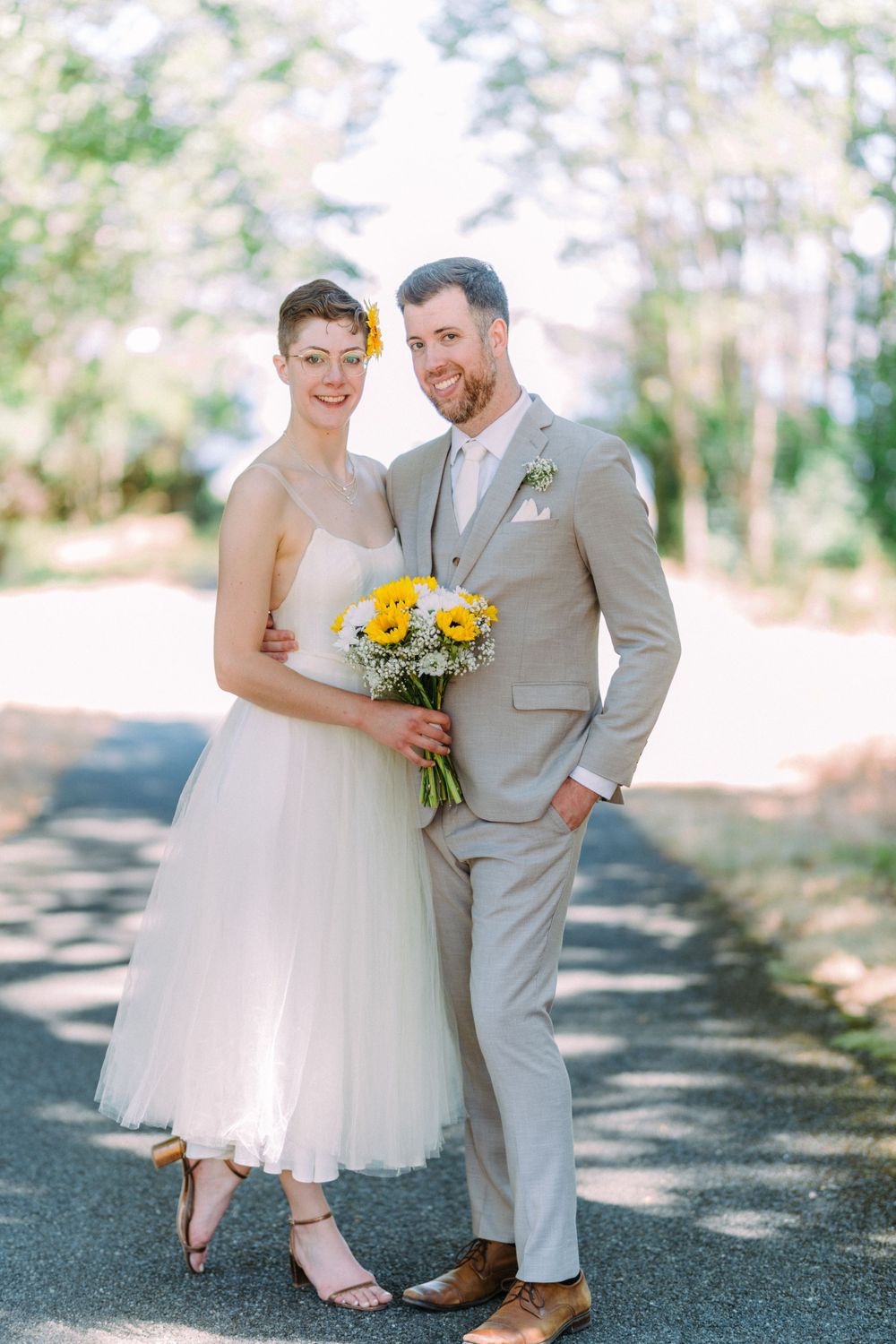 A joyful wedding couple poses outdoors, with the bride holding bright yellow sunflowers and wearing a tea-length dress.