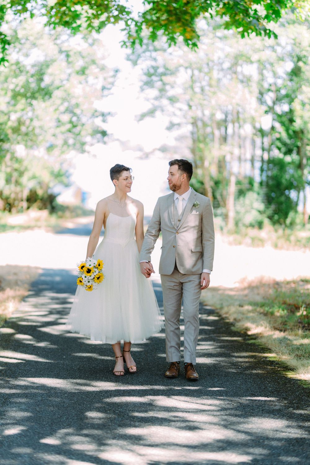 A wedding couple walks hand in hand down a tree-lined path with dappled sunlight.