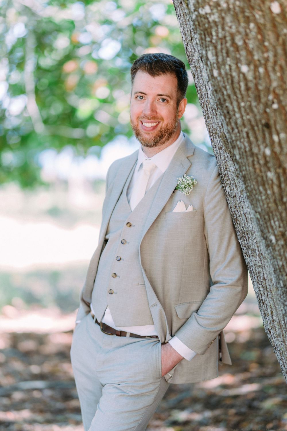 A light gray three-piece suit with boutonniere is showcased against a tree in natural lighting.