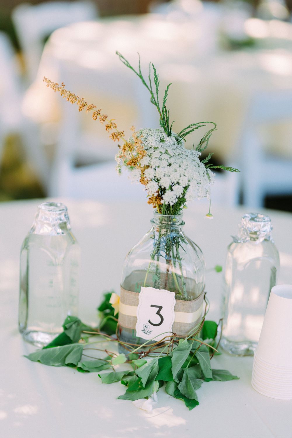 Rustic wedding centerpiece with white and yellow flowers in a glass jar vase marked with number 3.