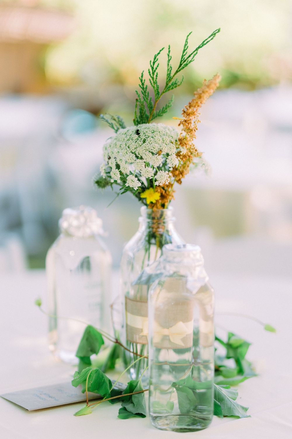Delicate floral centerpiece with white and yellow blooms in glass bottles surrounded by green leaves.