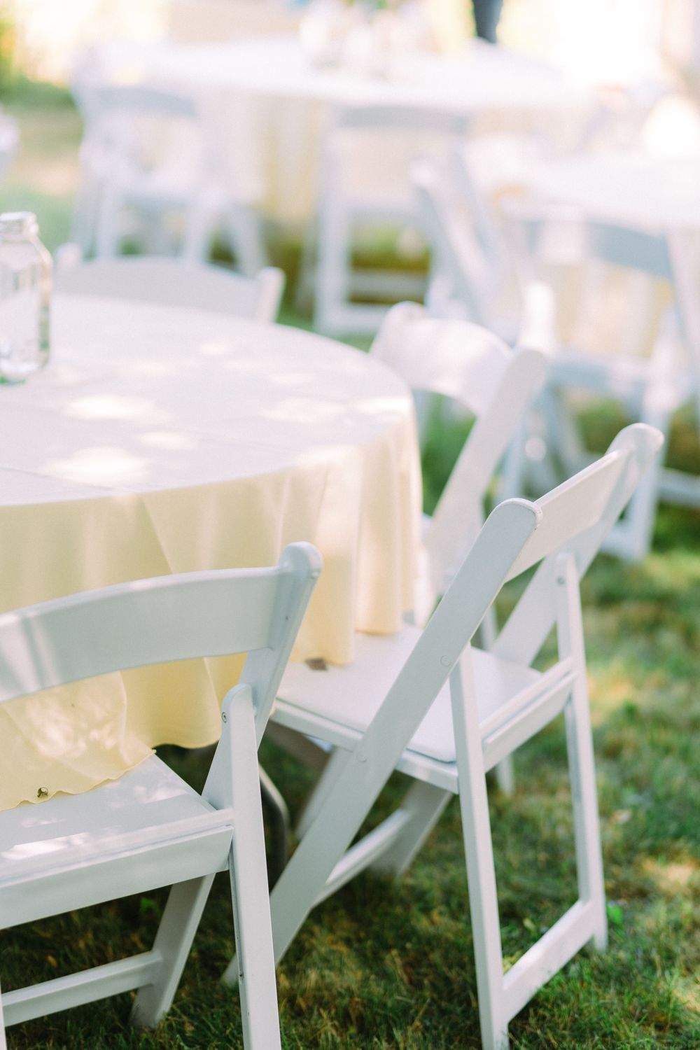 White folding chairs arranged around tables with cream tablecloths for outdoor wedding reception.