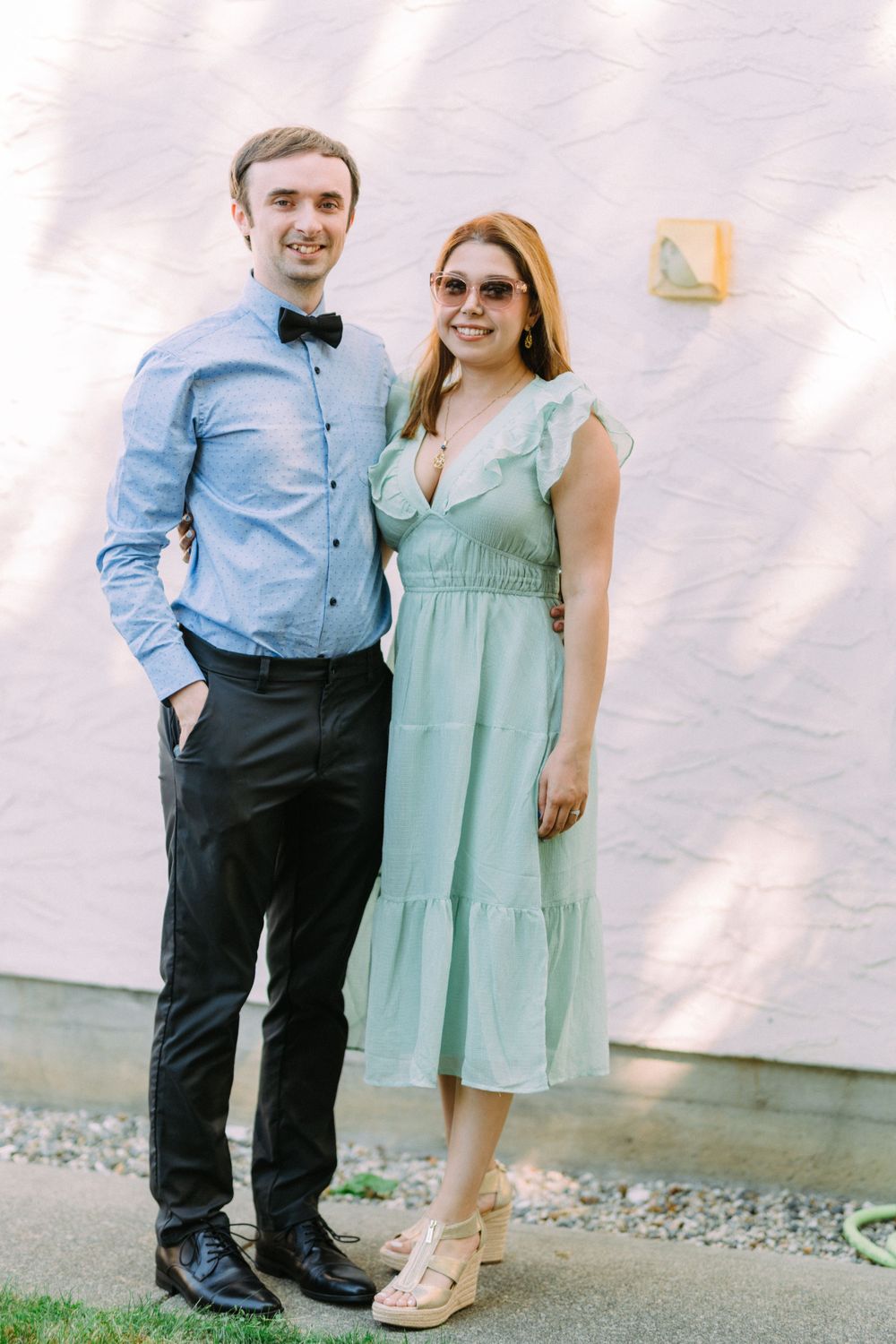 A couple dressed in formal attire posing against a light pink wall.