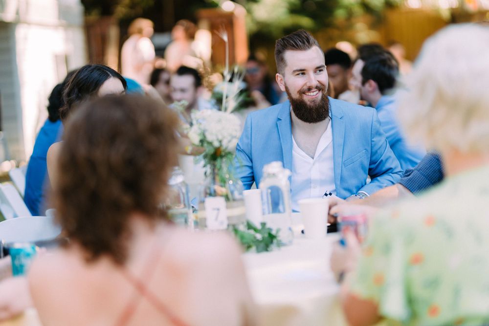 A guest in a light blue blazer enjoying conversation at an outdoor wedding reception.