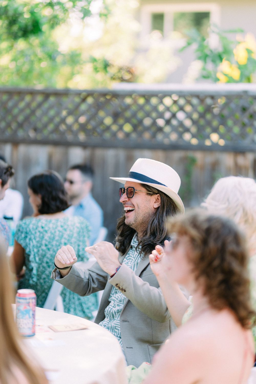 A guest wearing a panama hat and sunglasses animated in conversation at an outdoor party.