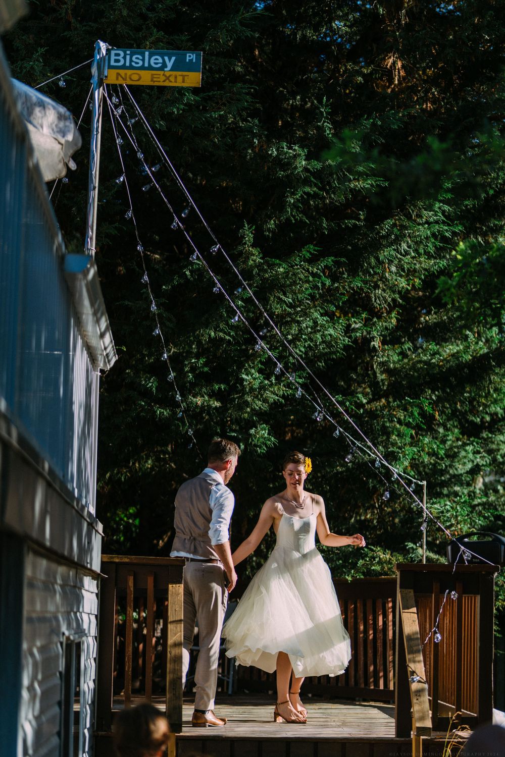 A couple dances together on a wooden deck beneath a street sign reading 'Bisley'.