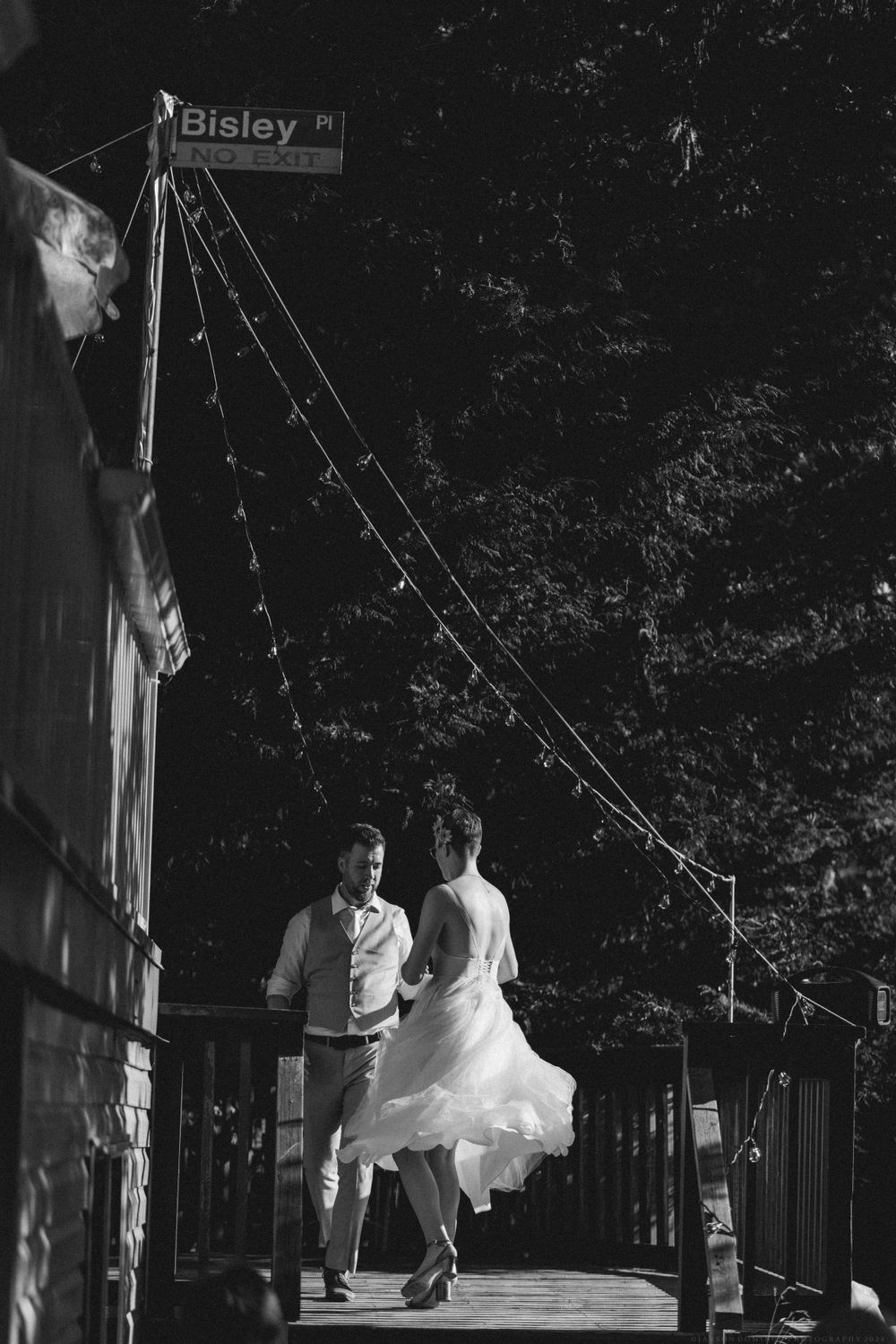 A black and white photo of a couple dancing on a deck under strung lights and a Bisley street sign.