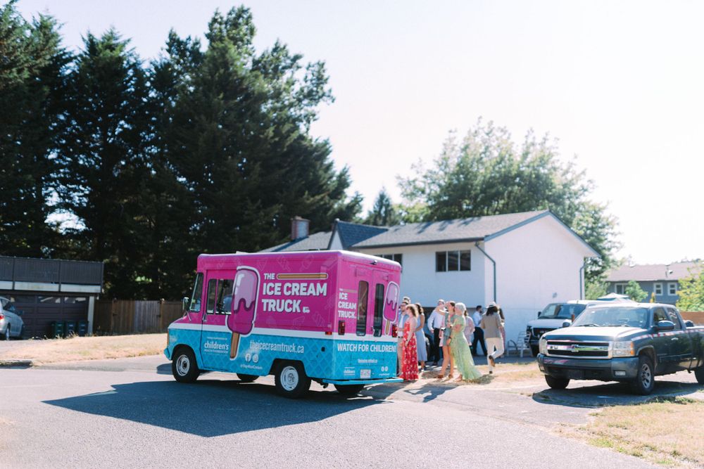 A bright pink ice cream truck parked outside a residential area with customers waiting.