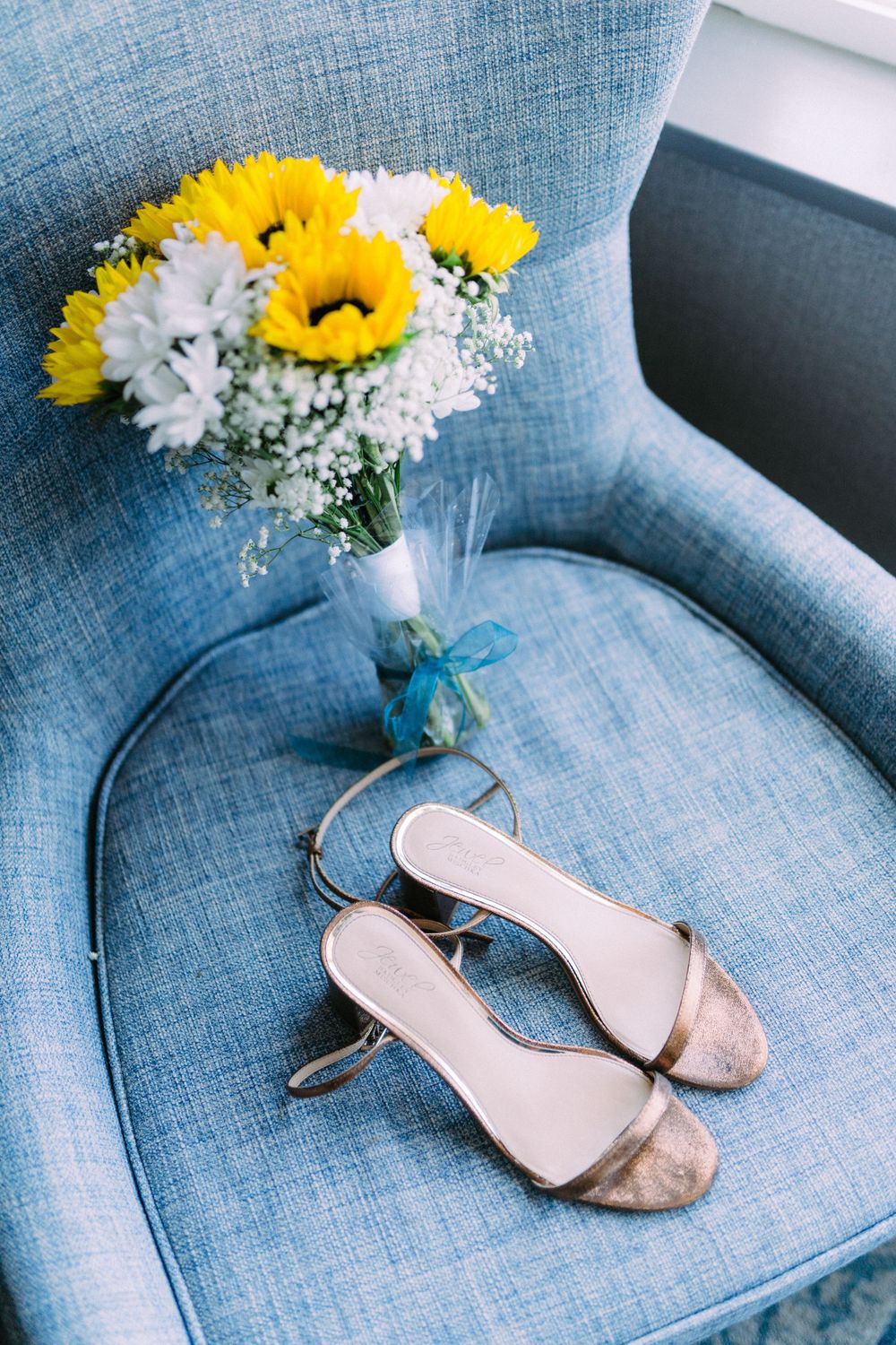 Wedding shoes and sunflower bouquet arranged on light blue upholstered chair.