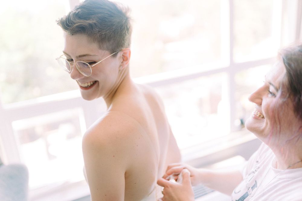 A person wearing glasses gets dressed in a sun-lit room while being helped by someone else.