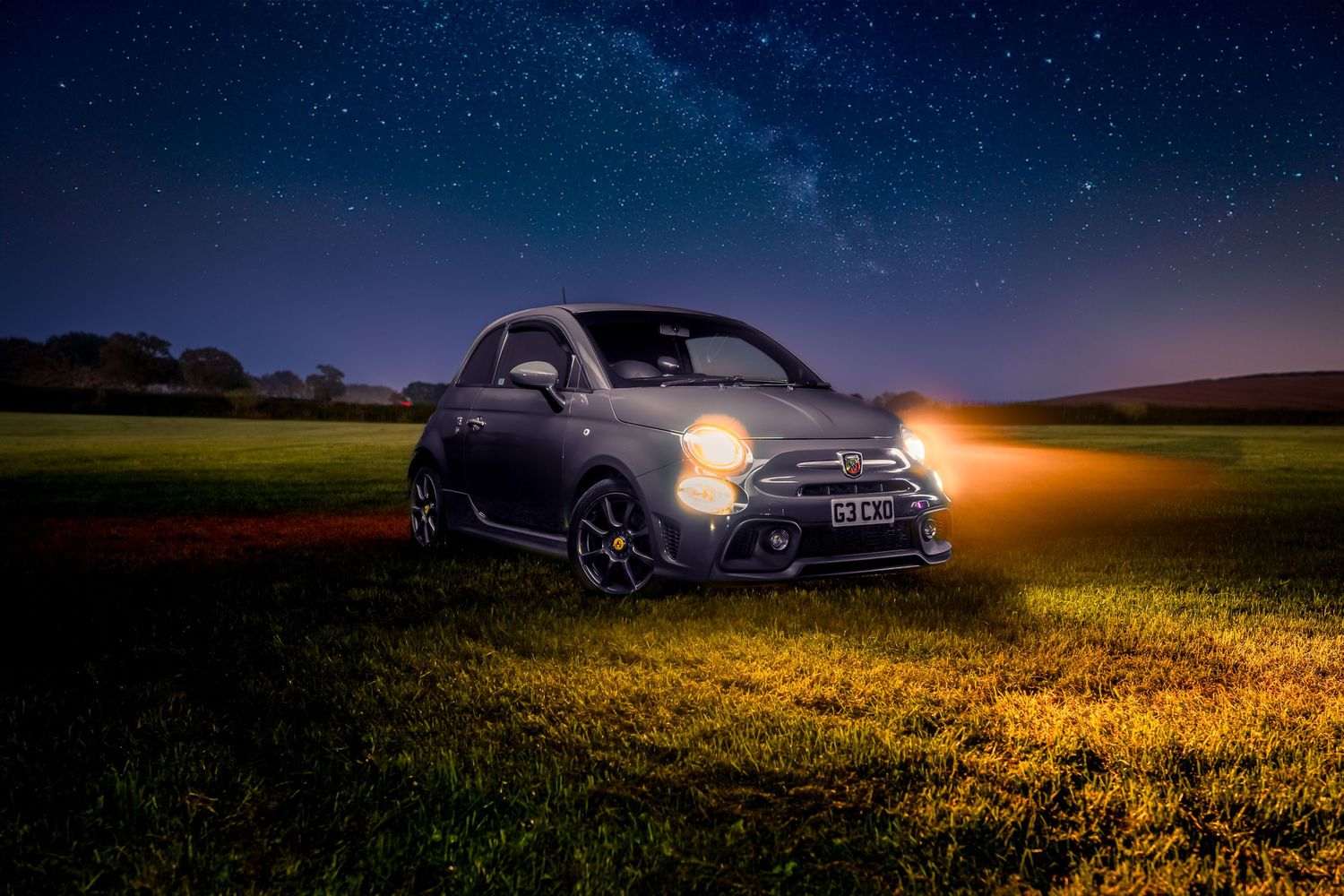 Fiat 500 car illuminated at night in a grassy field under starry sky with headlights glowing.