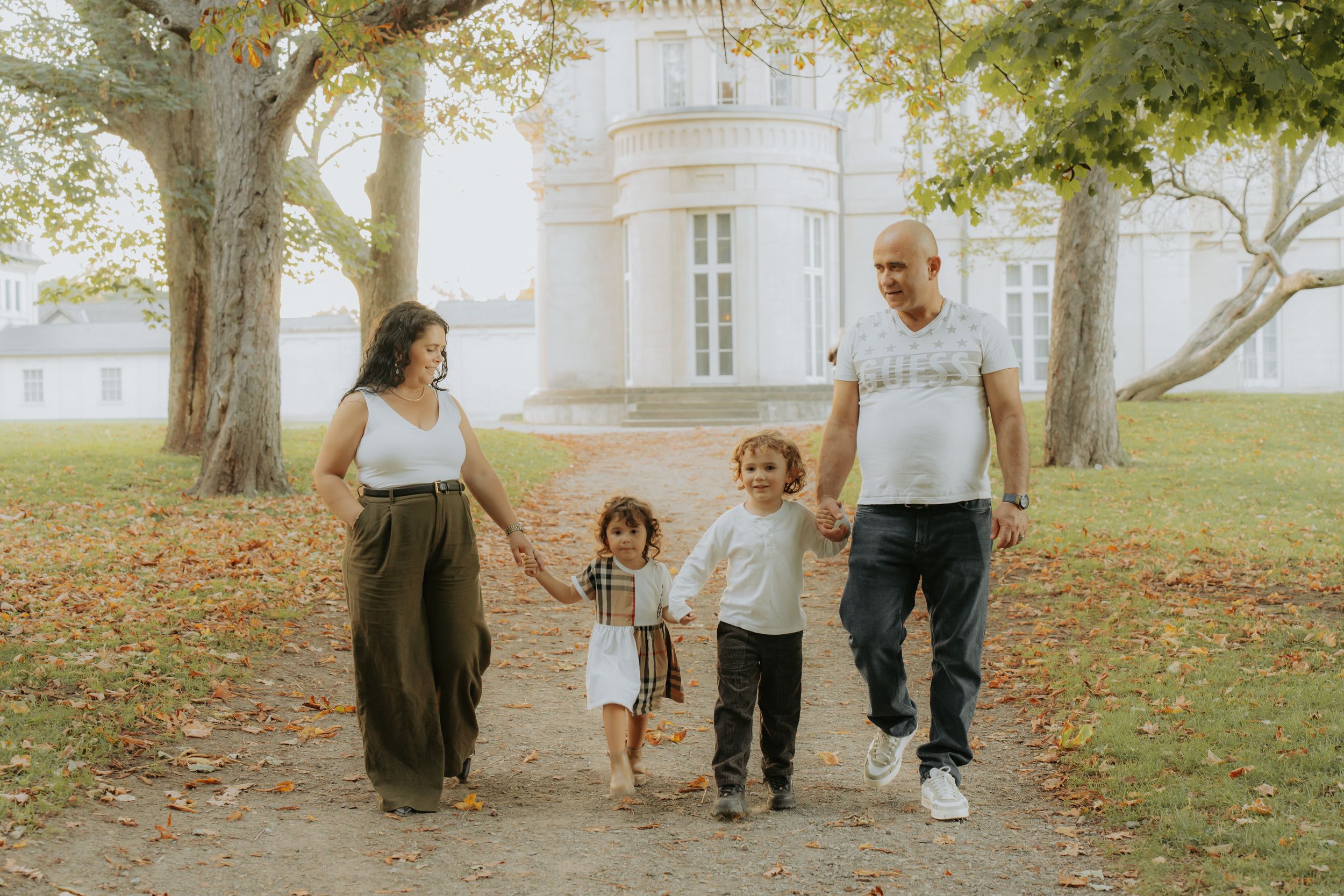 A family of four holding hands, walking down a tree-lined path in front of Dundurn Castle