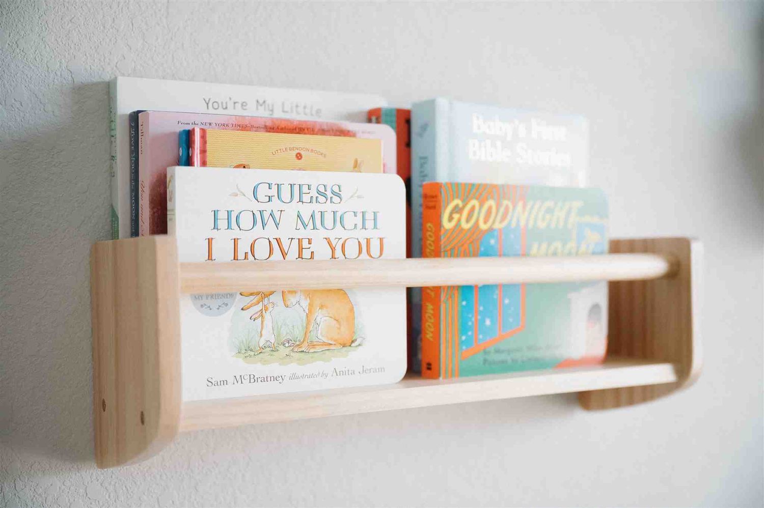 Children's books displayed on a minimalist wooden wall-mounted shelf, featuring 'Guess How Much I Love You' and other titles.