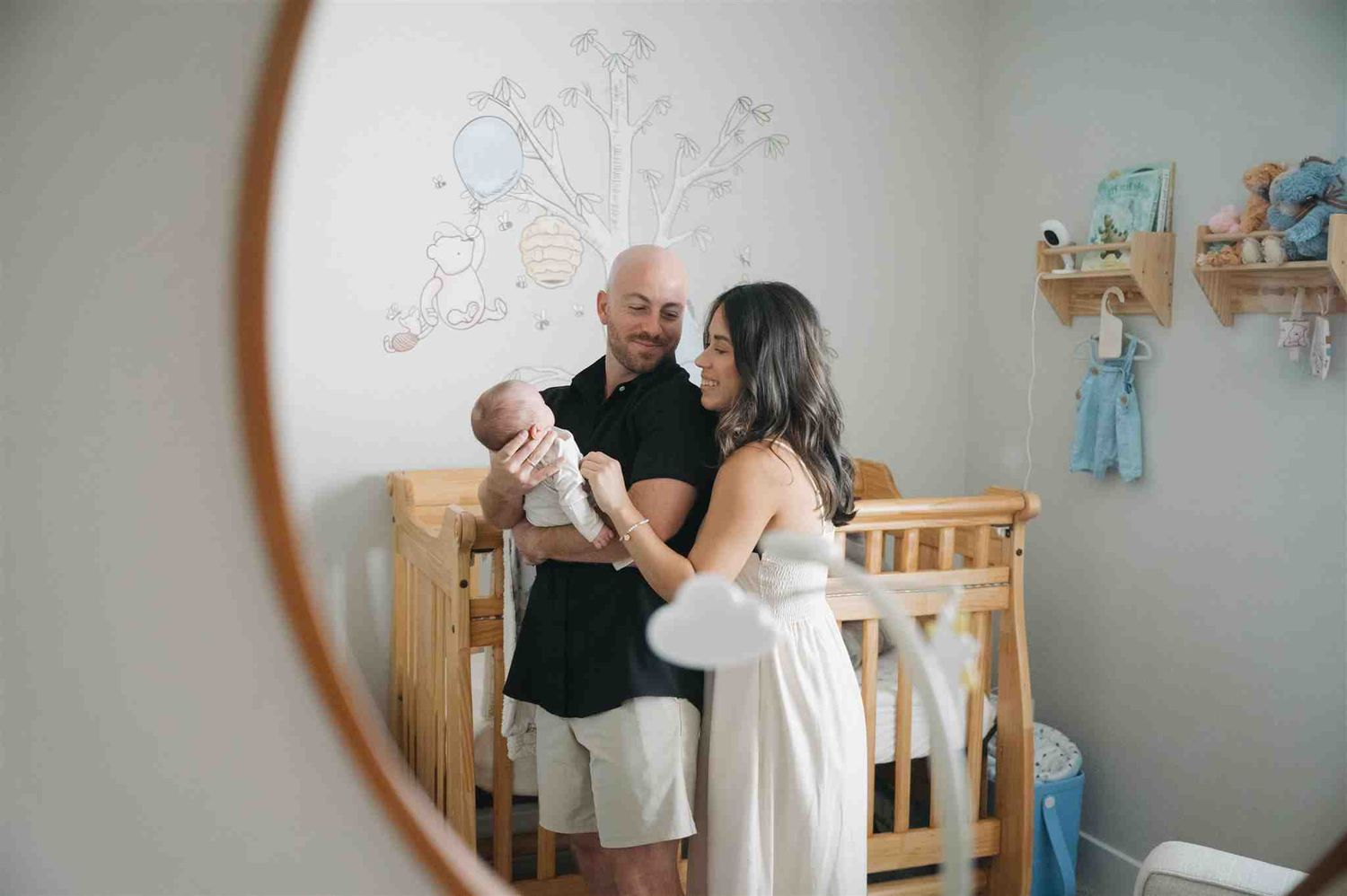 Two people with their infant stand near a wooden crib in a nursery with wall decor visible through a circular mirror frame.