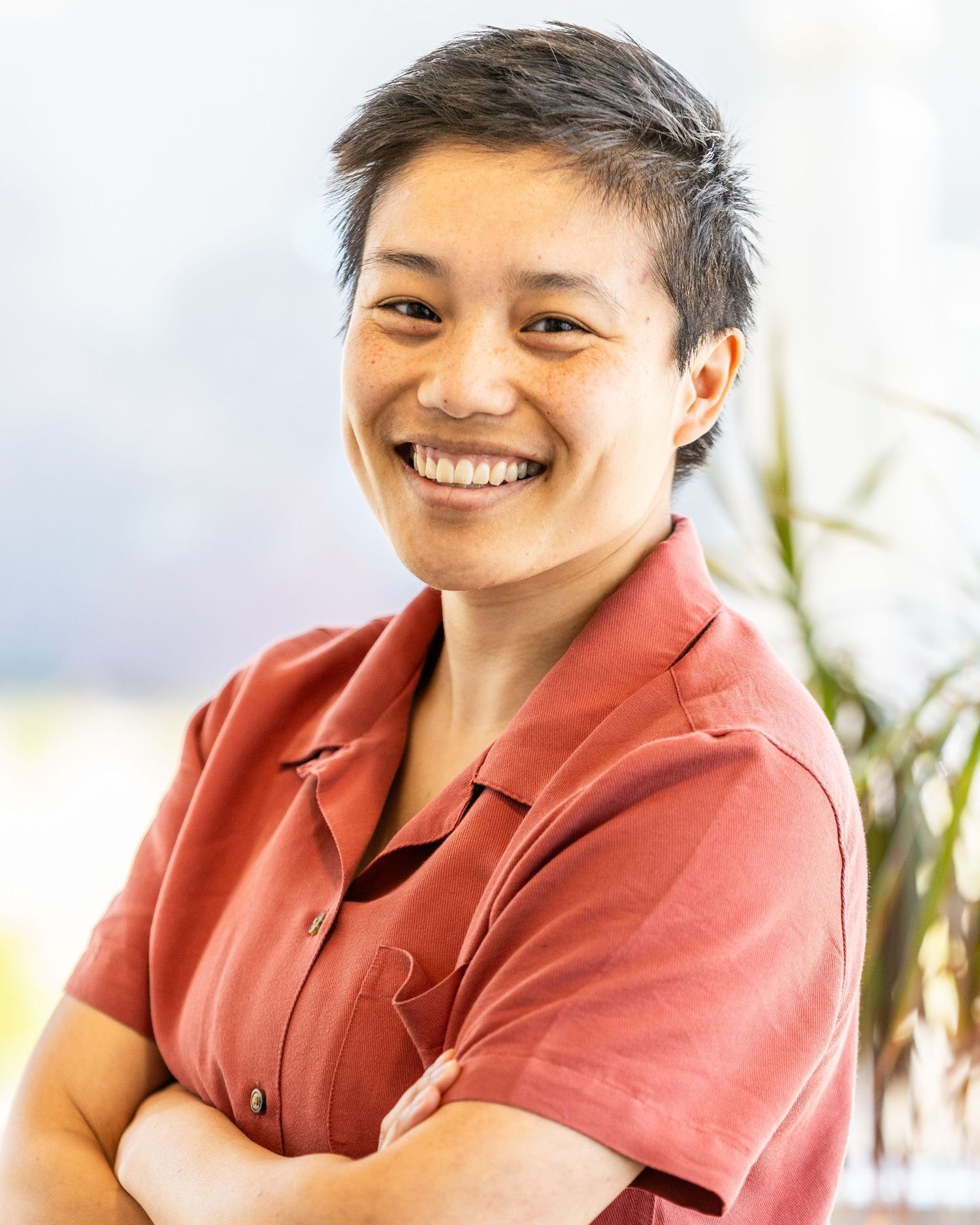 Professional headshot of a person in a coral button-up shirt smiling warmly with crossed arms against a bright background.