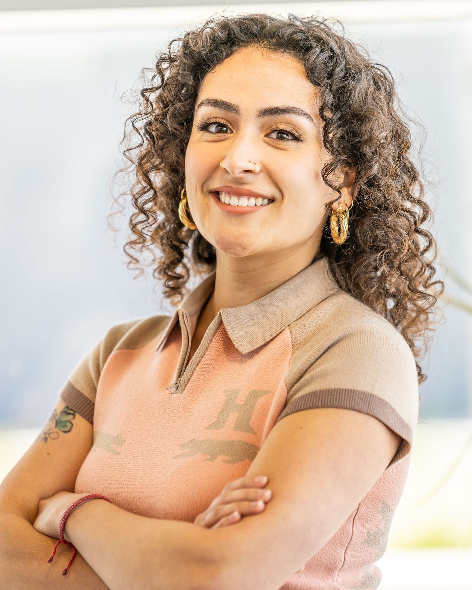 Professional Headshot of someone with natural curly hair wearing a beige collared shirt, smiling warmly with arms crossed.