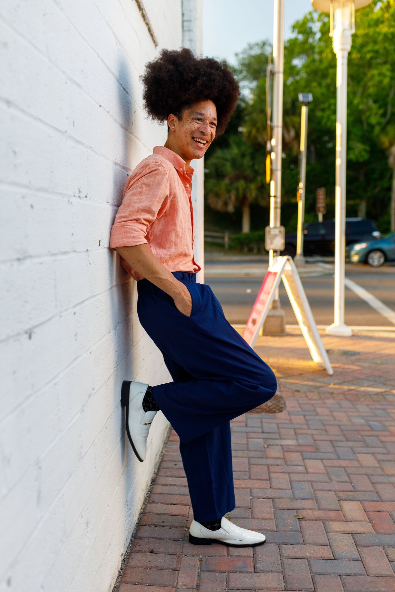 Jeremiah in coral button-down shirt and navy pants leans against white wall on brick sidewalk, smiling outdoors.