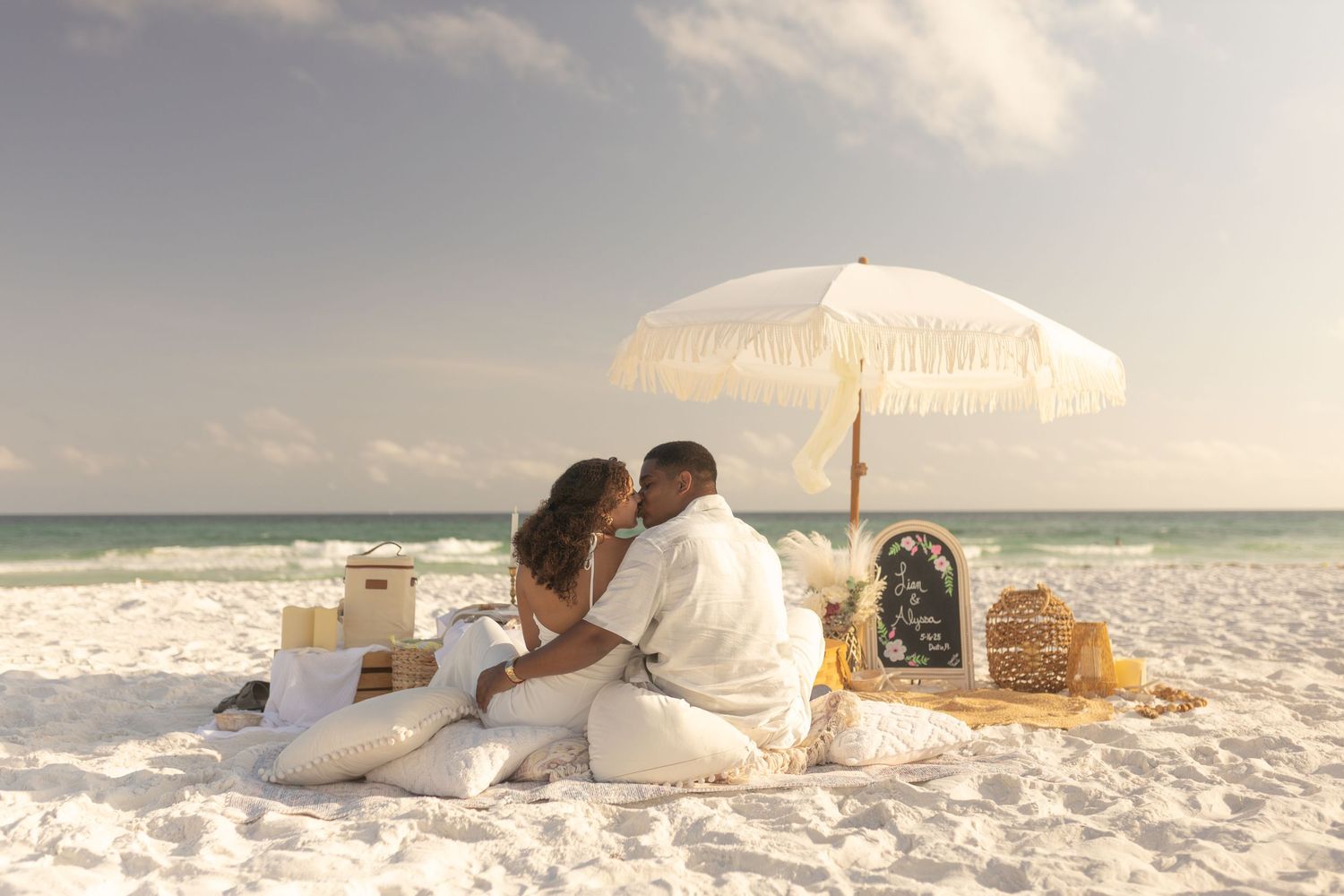 A romantic beach picnic setup with a white umbrella, wicker baskets and blanket on pristine white sand by turquoise waters.