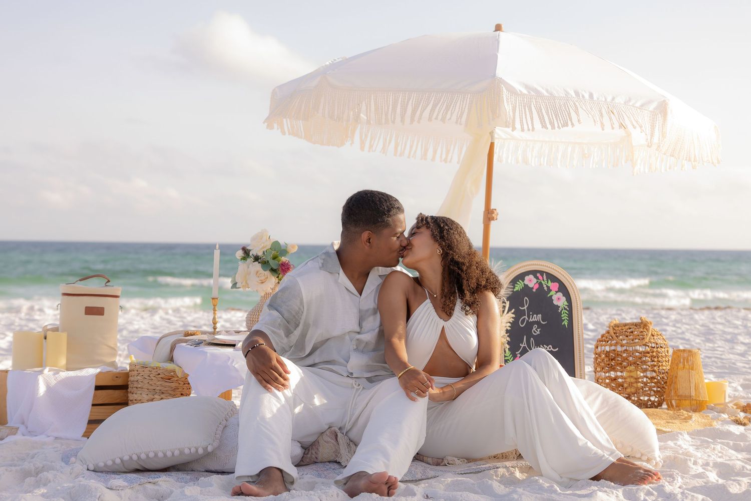 A romantic beach picnic setup with a white parasol, lanterns and wicker baskets on the sand by the ocean.