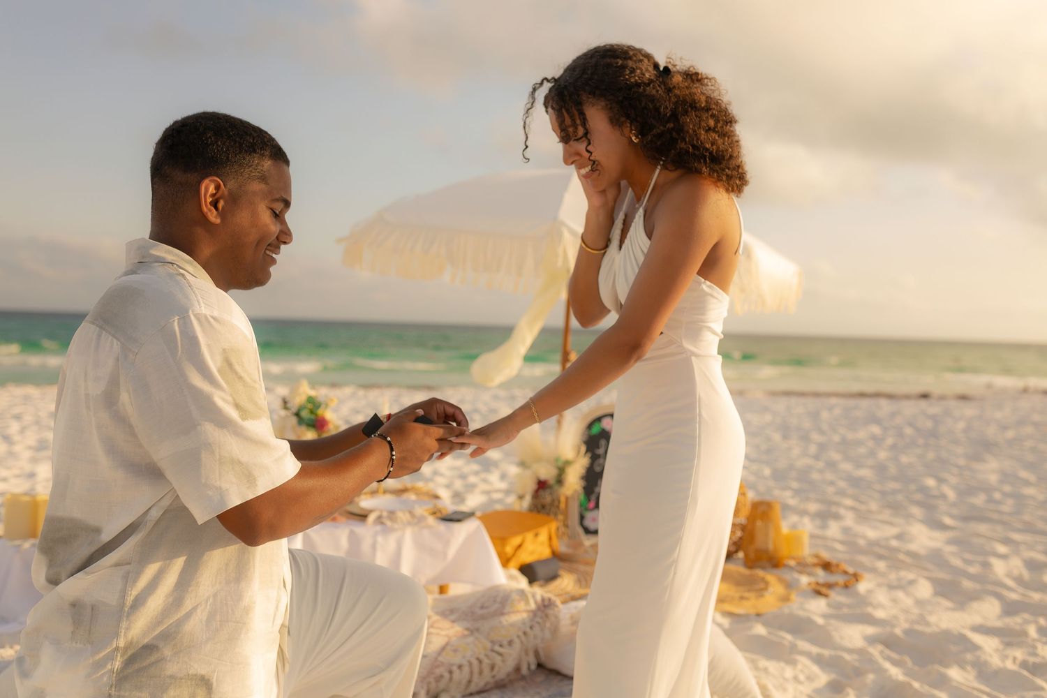 A couple shares a romantic proposal moment on a beach at sunset with yellow flowers in the sand.