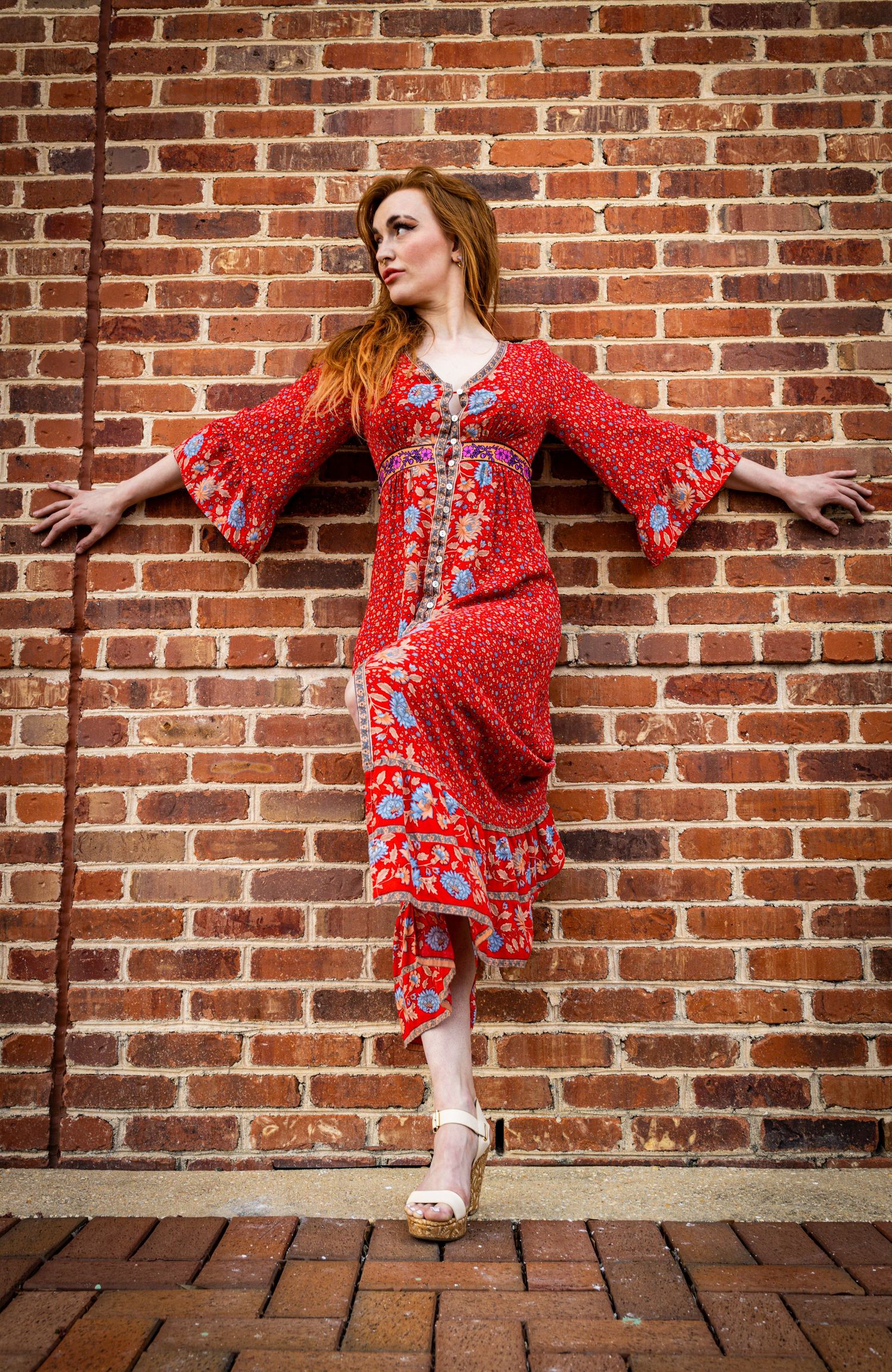 Woman in flowing red patterned dress poses expressively against rustic brick wall.