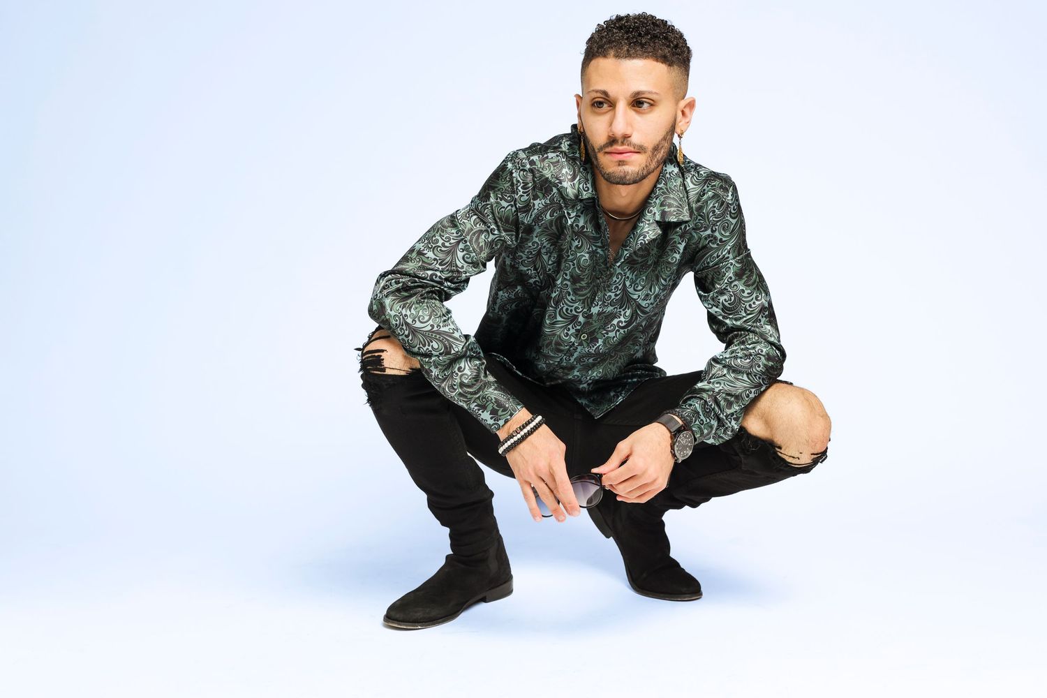 Person in patterned jacket and ripped jeans crouches against white background.