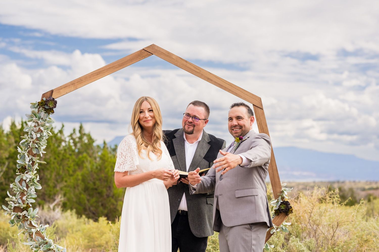 A wedding ceremony takes place under a wooden arch frame against a desert landscape with mountains.