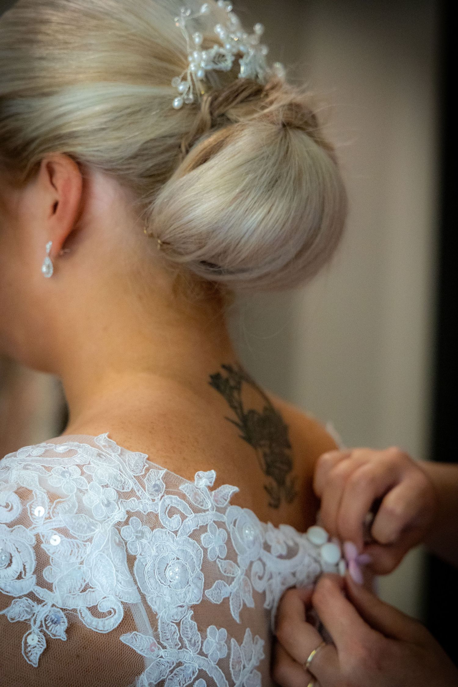 Close-up of bride's lace wedding dress being fastened, revealing floral shoulder tattoo.