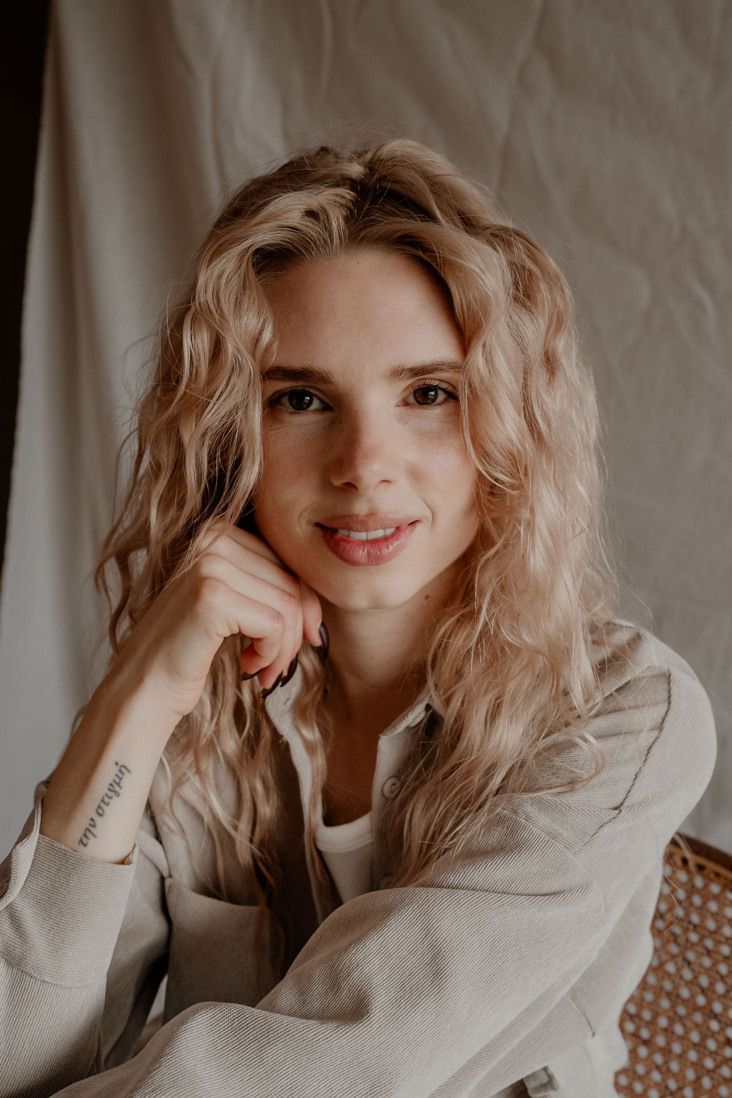 Portret van een persoon met golvend blond haar, gekleed in een beige trui, met de hand rustend op de kin in warm licht.