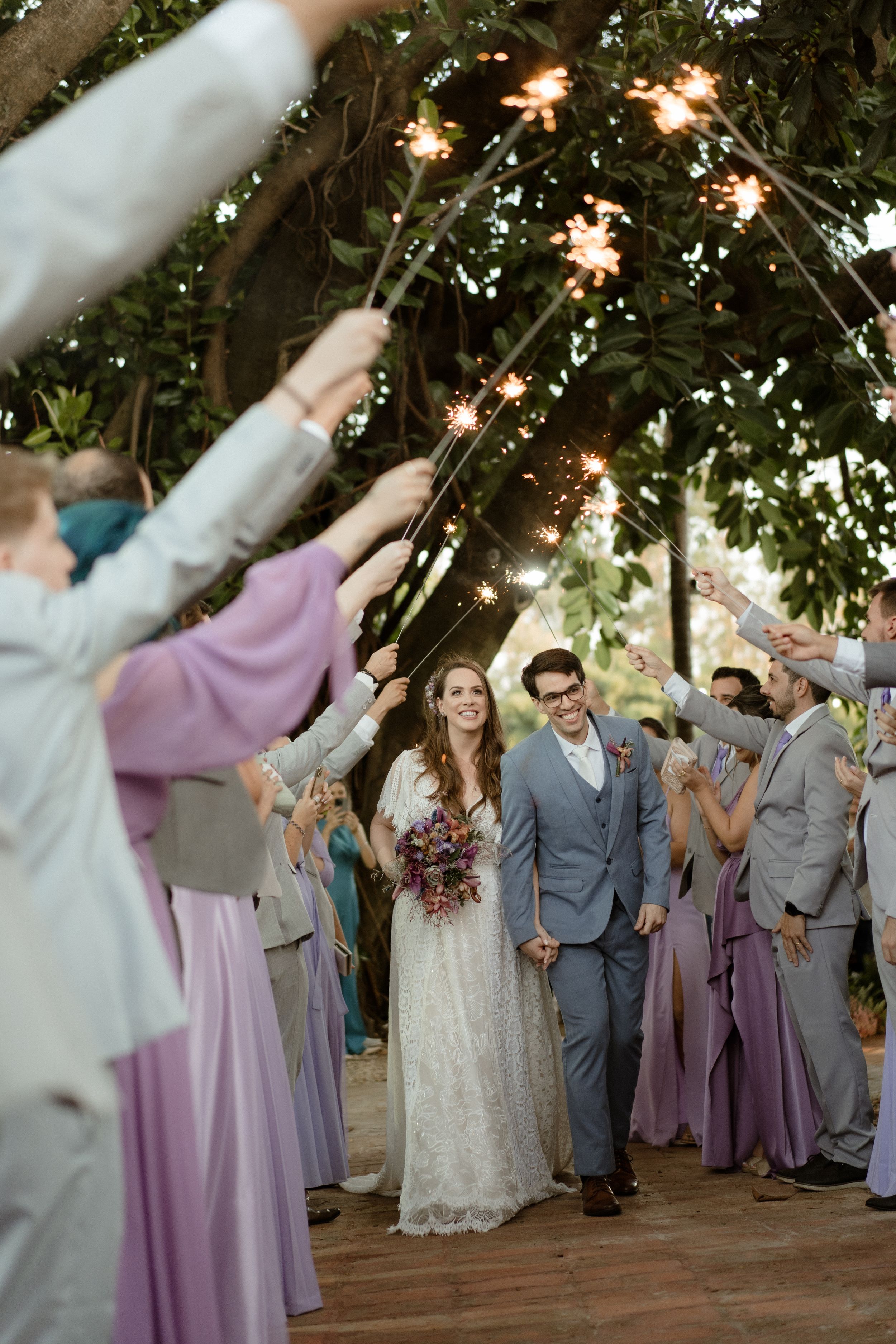 Wedding party celebrates with arms raised under twinkling lights and tree canopy.