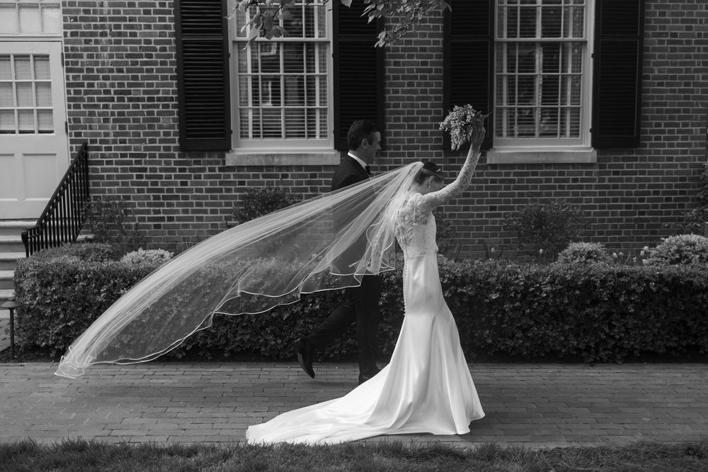 A black and white photo of a bride in a flowing gown with veil billowing outside a brick building.