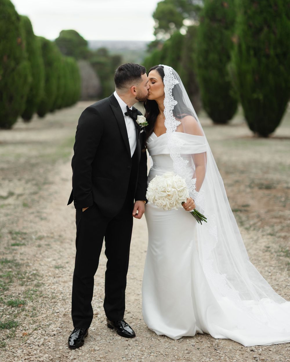Bride and groom sharing a romantic kiss on a tree-lined path, bride in white off-shoulder gown with veil and bouquet.