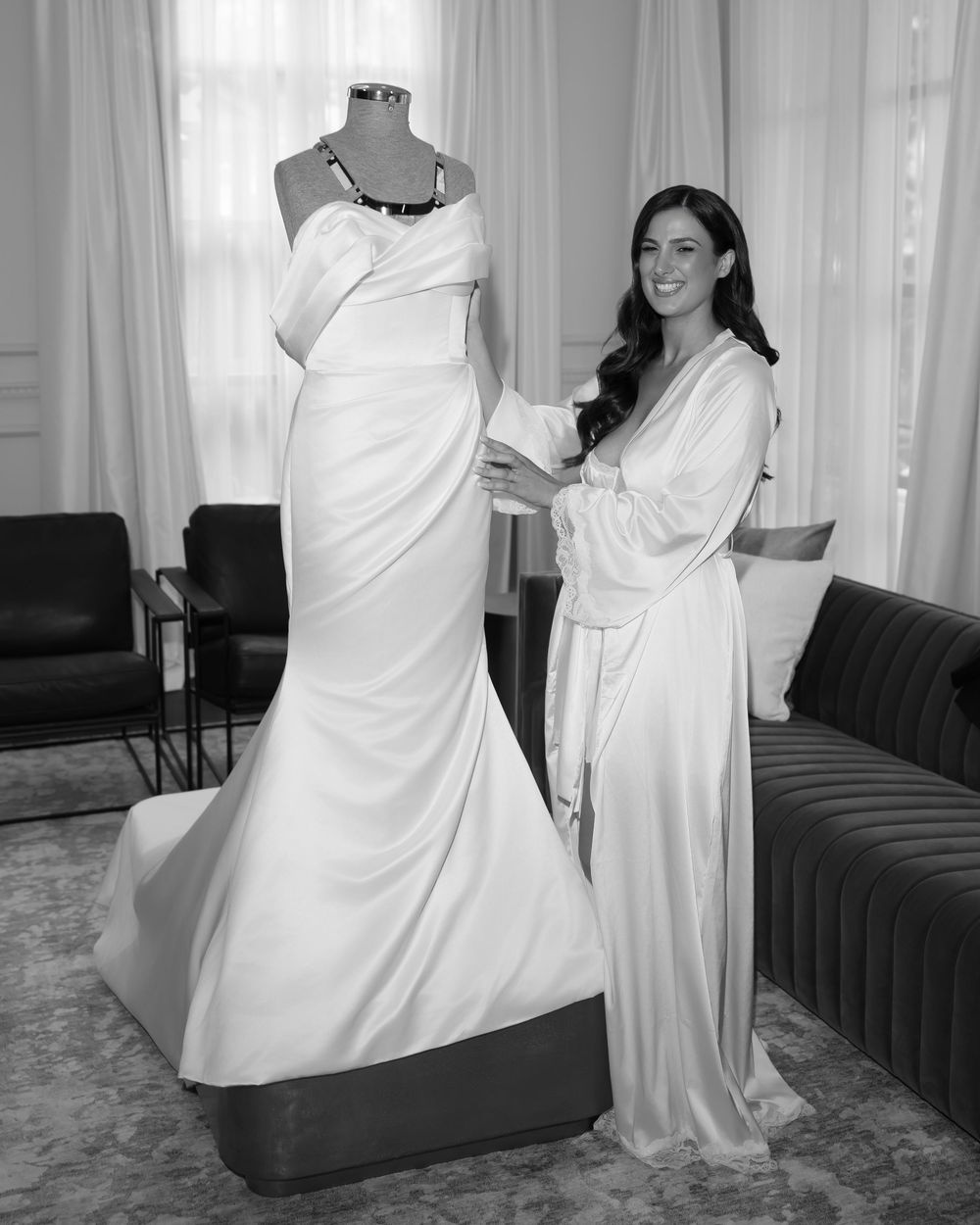 Woman in robe poses beside elegant mermaid wedding gown on mannequin in black and white photo.