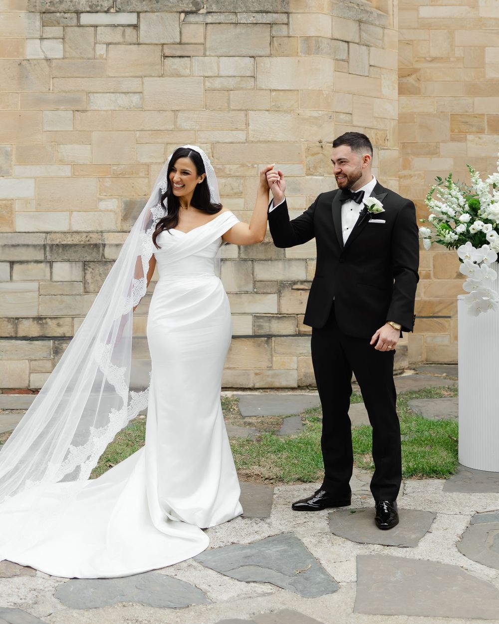 Bride in white off-shoulder gown and veil holding hands with groom in black tuxedo at outdoor ceremony.