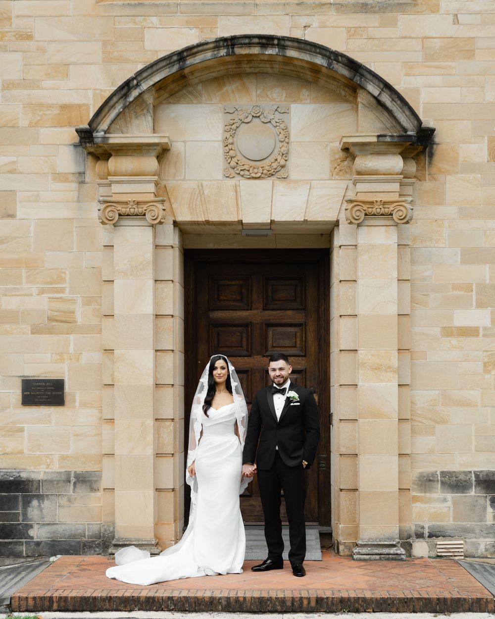Bride and groom posing hand in hand before an ornate sandstone church doorway, bride in white gown with veil.
