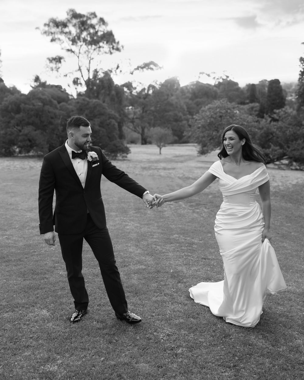Black and white photo of a bride in a white gown and groom in a tuxedo holding hands on a lush green lawn.