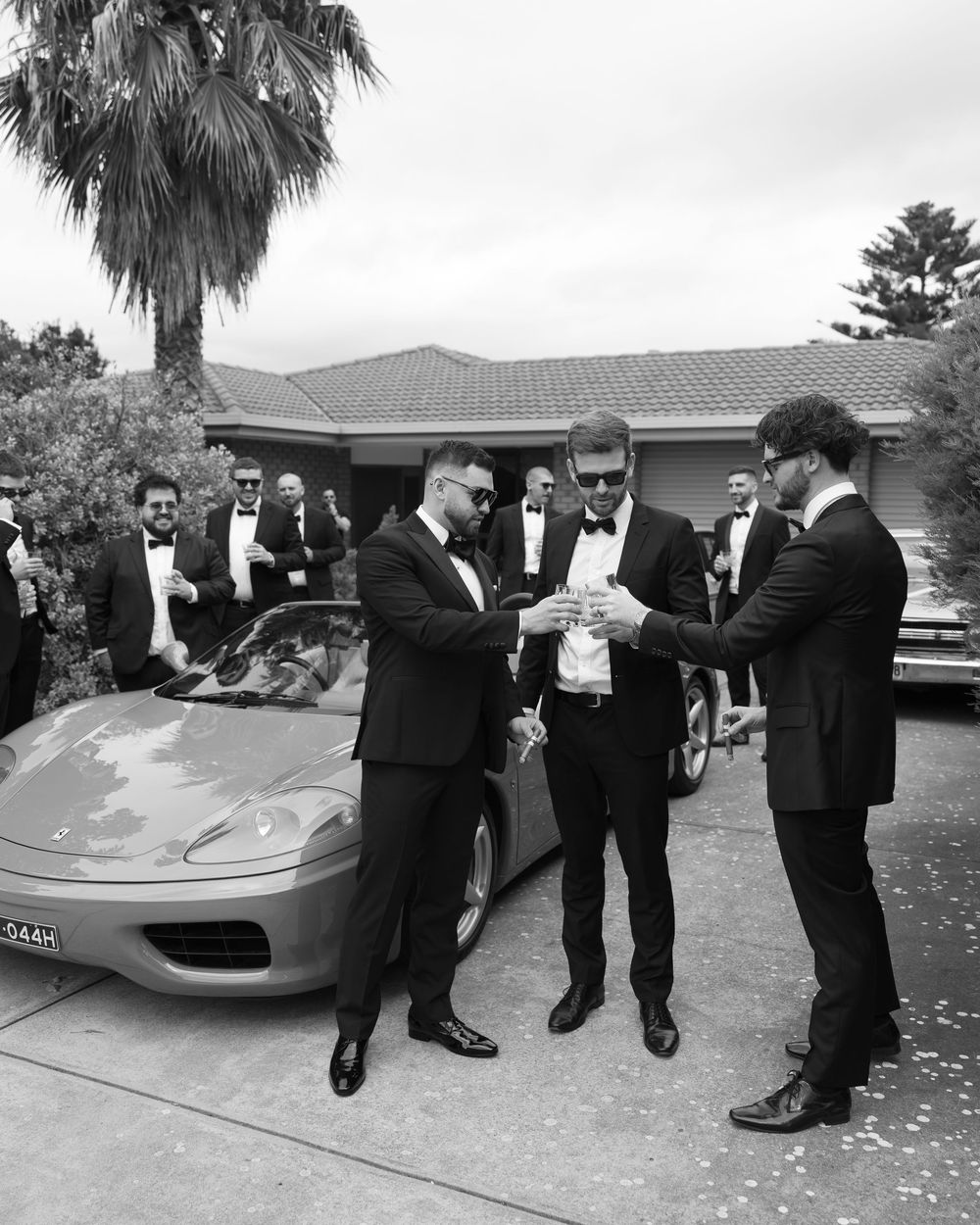 Groomsmen in black tuxedos toasting beside a Ferrari sports car outside a home before a wedding.
