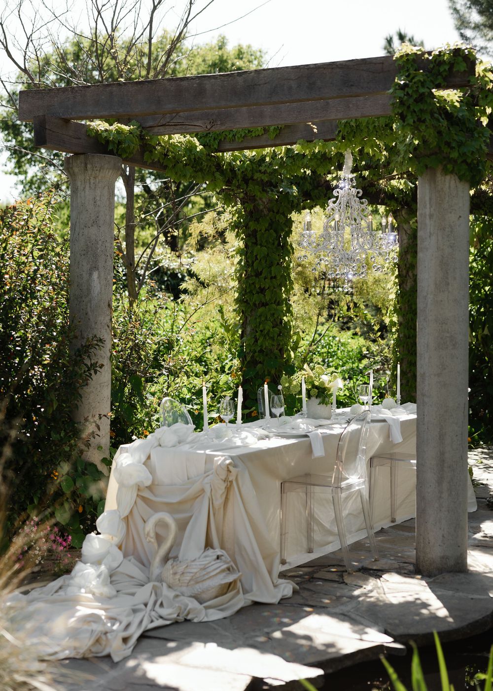 Elegant outdoor dining setup under a vine-covered pergola with white tablecloths and chair covers.