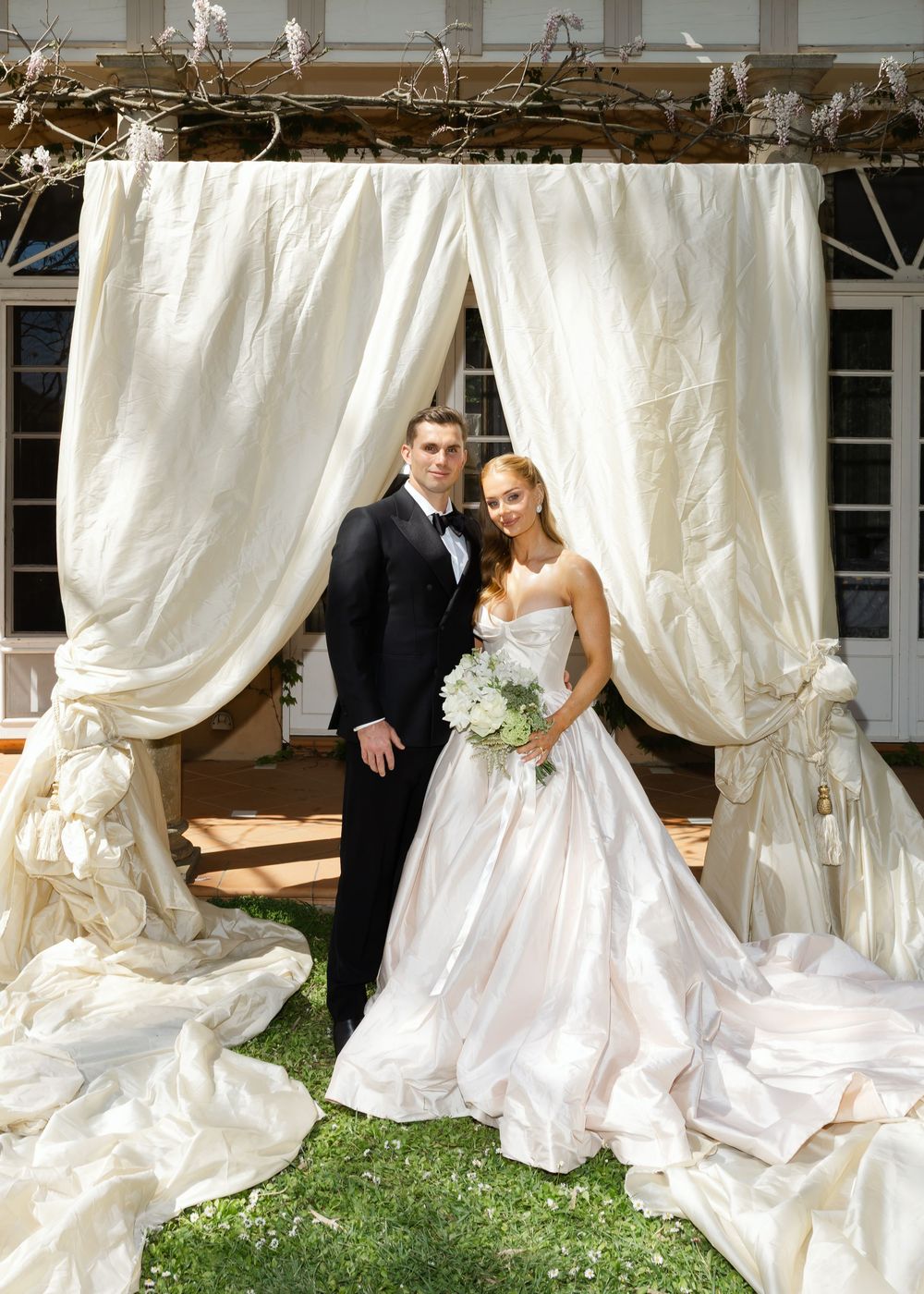 Wedding couple poses beneath flowing white fabric drapes in an outdoor garden setting.